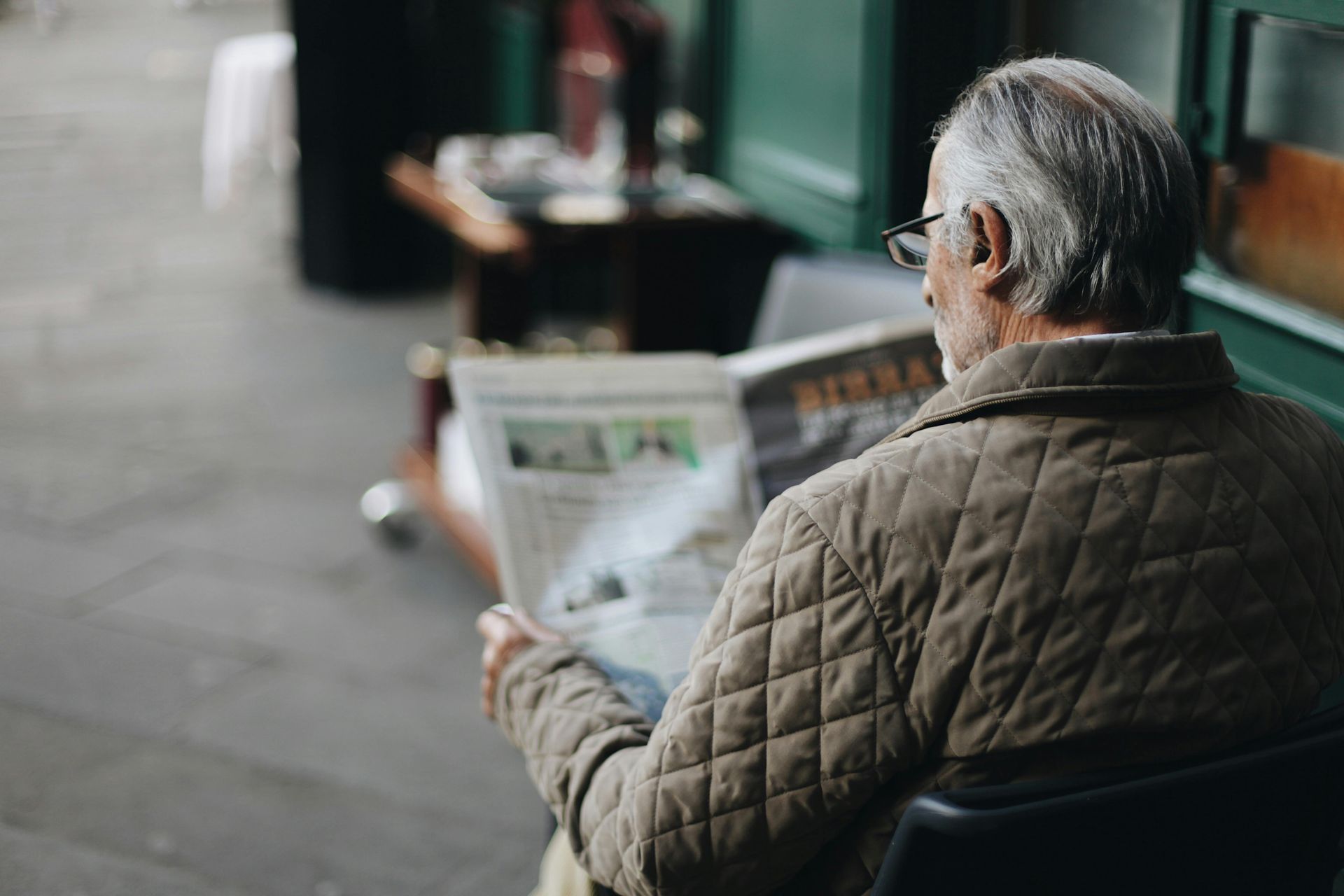 Man reading a newspaper outdoors, wearing glasses and a quilted jacket.