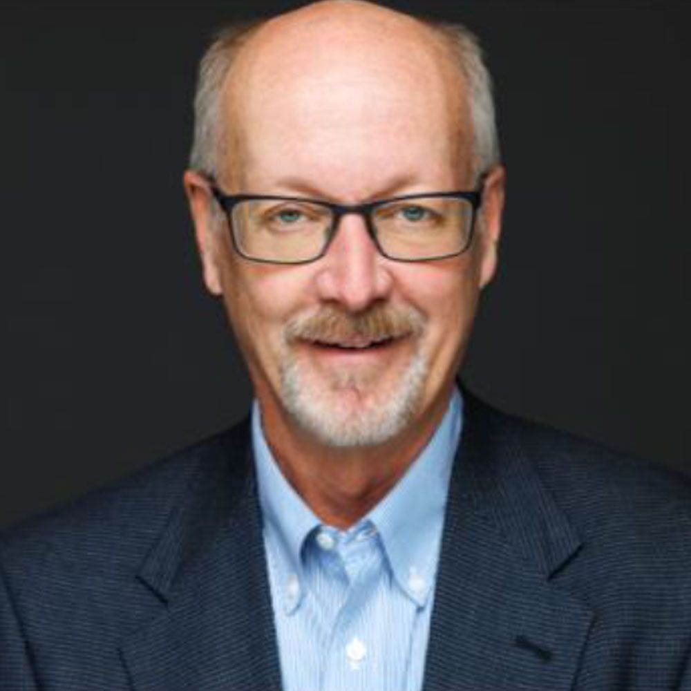 Man with glasses, wearing a blue button-down shirt under a navy blazer, smiling at the camera.
