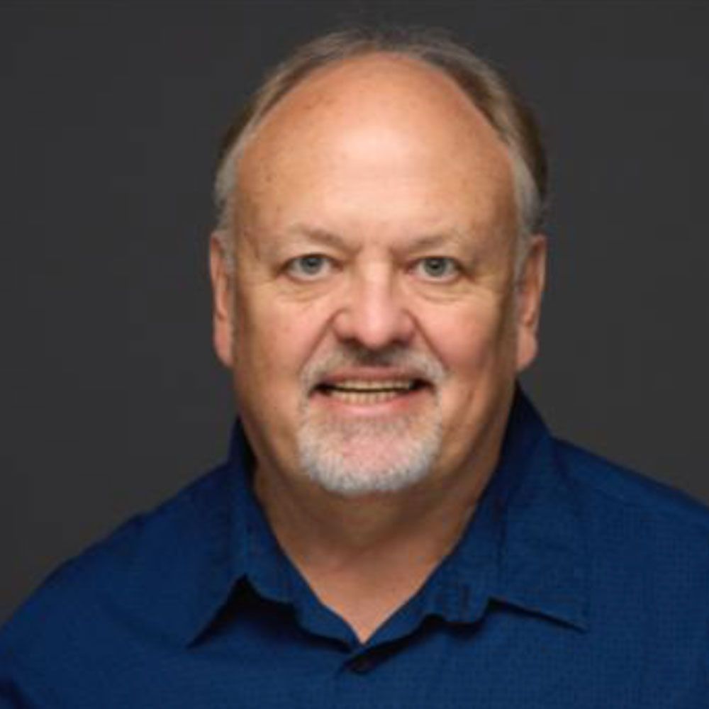 Man in blue shirt smiling at the camera, short gray beard, neutral gray backdrop.