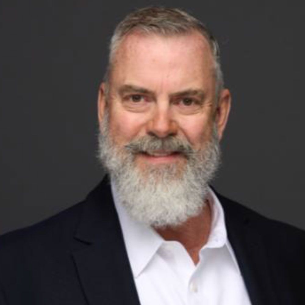 Man with gray beard smiles, wearing a navy suit jacket and white shirt.