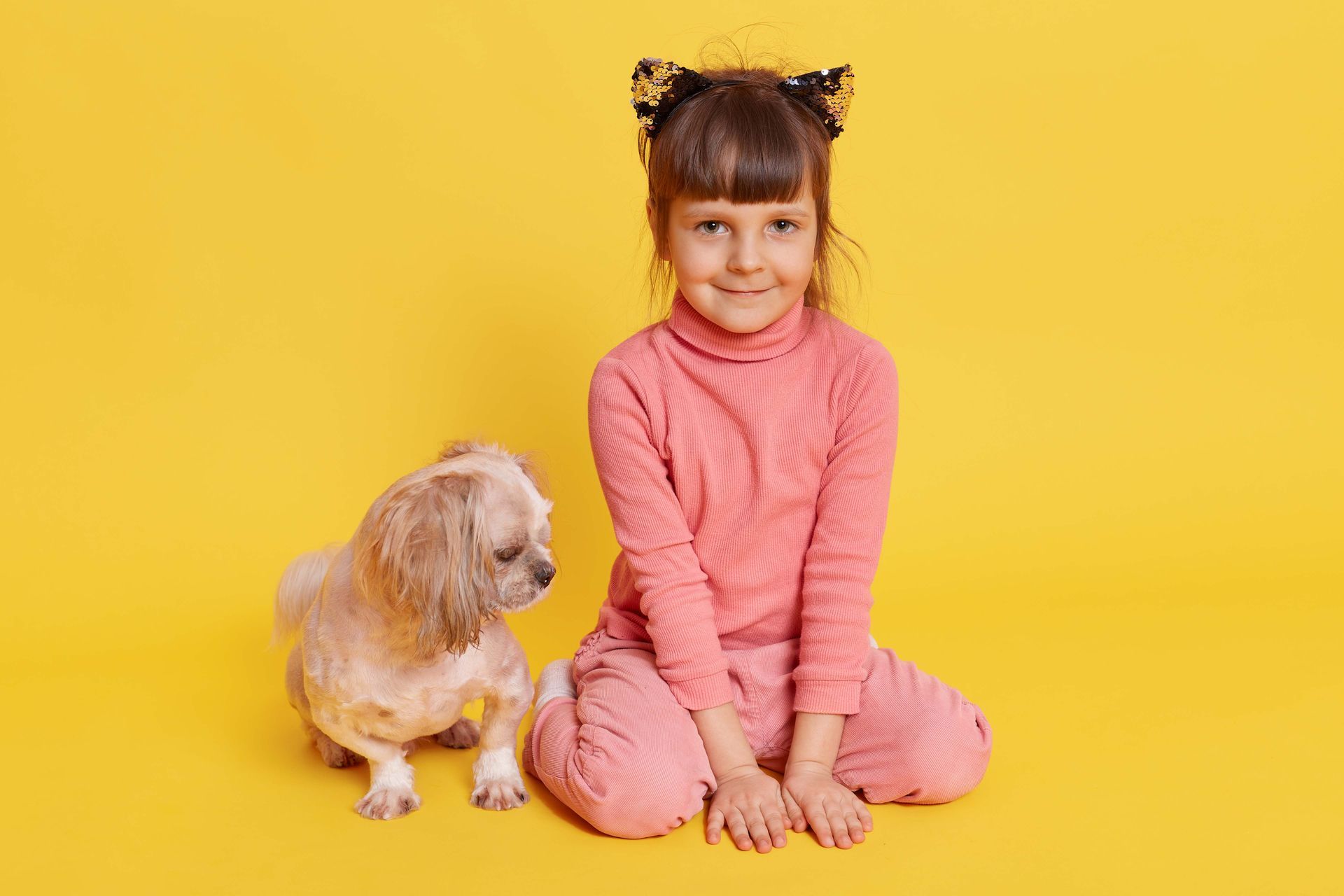 A child with cat ear hair accessories sits on a yellow background next to a small, light-colored dog.