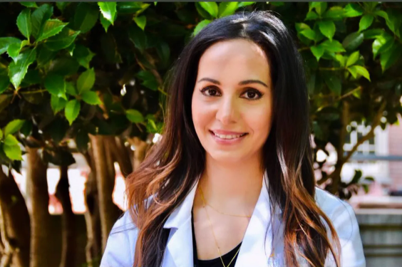 A smiling woman with long, dark hair in a white coat stands in front of greenery, likely outside.
