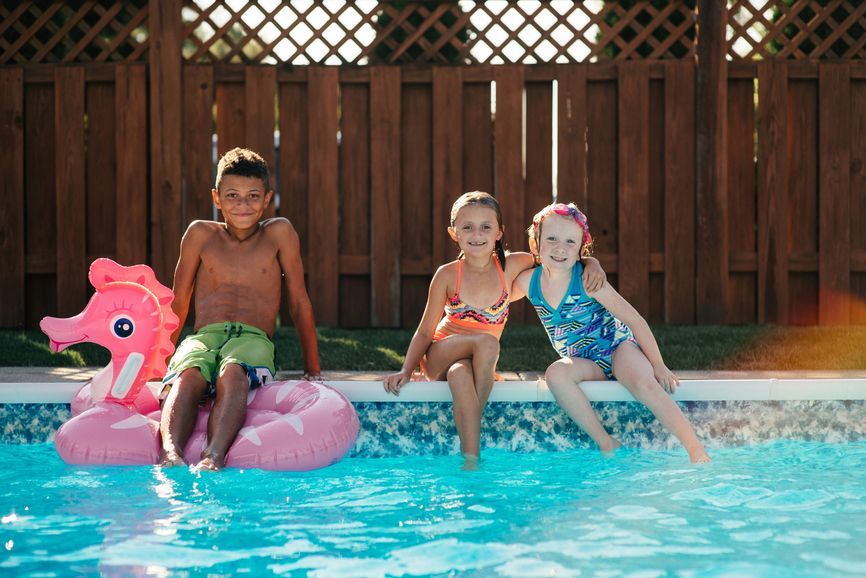 Three children sit on the edge of a pool, feet in the water. A boy sits on a pink seahorse float. Wooden fence in the background.