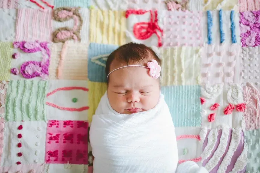 Newborn baby swaddled in white on a patchwork quilt, wearing a pink floral headband.
