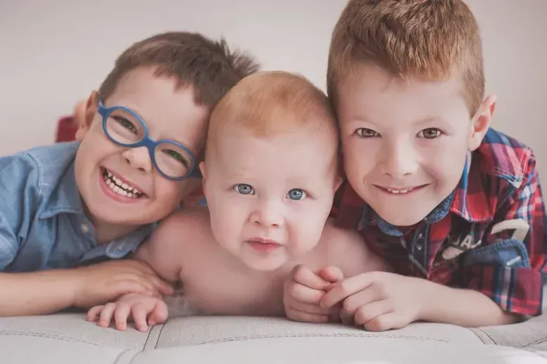Three smiling children, one baby and two older boys, lying close together, with a neutral background.