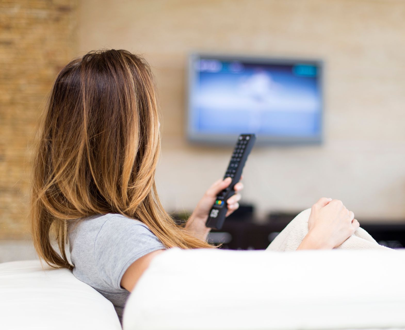 A woman is sitting on a couch holding a remote control in front of a television.