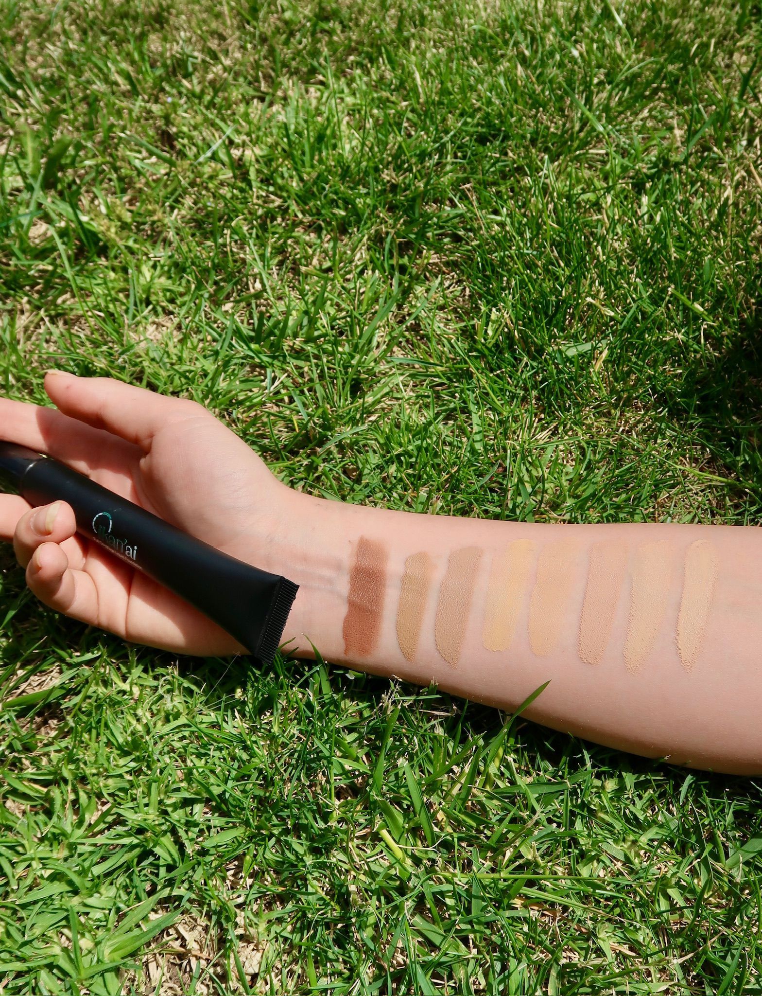 Arm with makeup swatches on grass, holding a black tube of product. Brown and gold tones  — Ikan'ai In Bowral, NSW