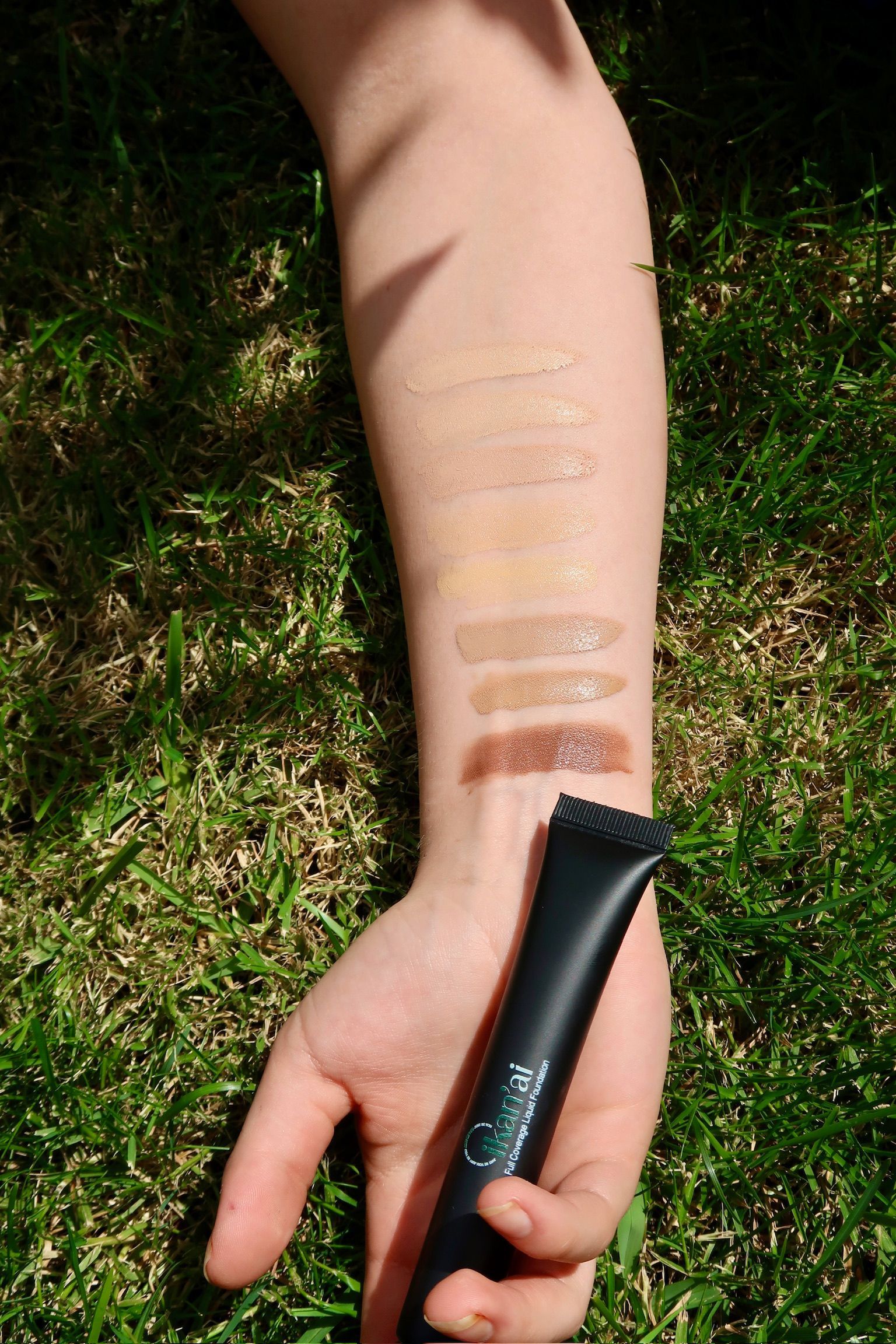 Arm with foundation swatches in various shades, next to a black tube, on green grass  — Ikan'ai In Bowral, NSW