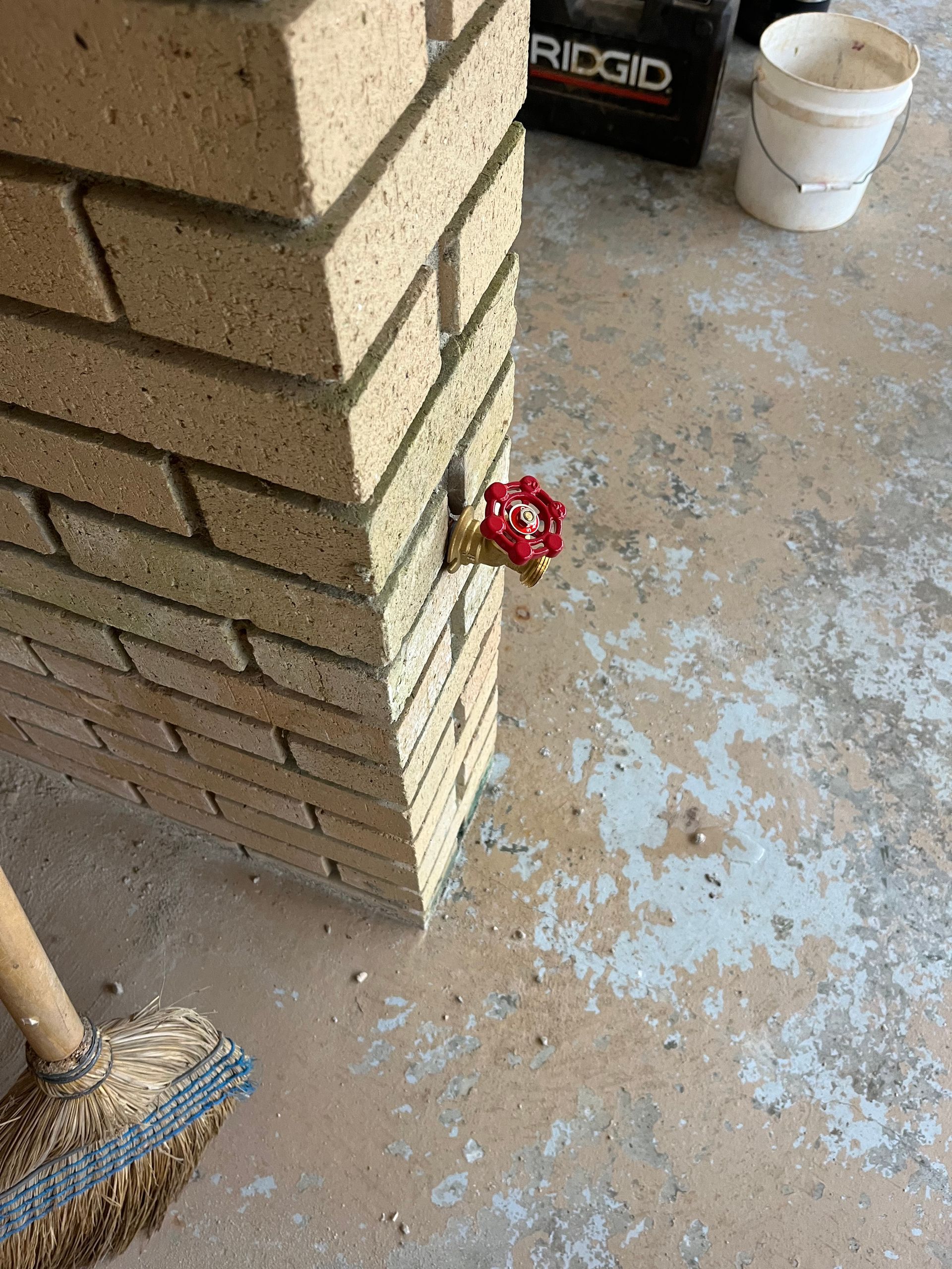 A broom is sitting next to a brick wall with a red valve on it.