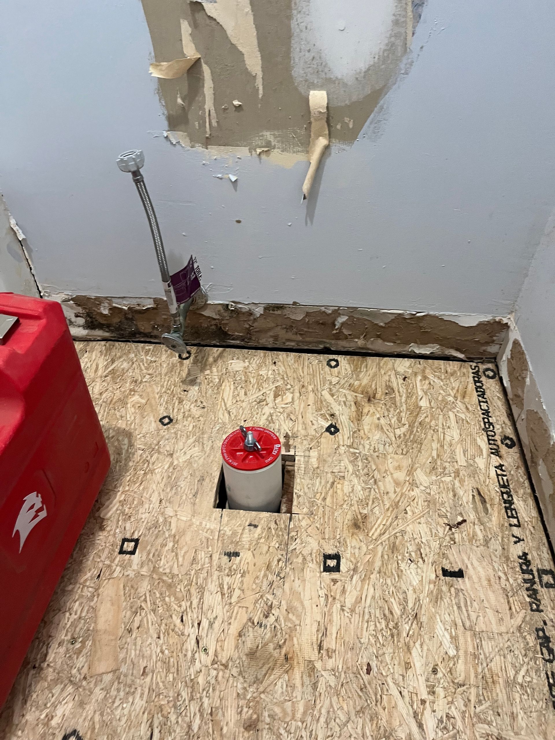 A red box is sitting on a wooden floor next to a hole in the wall.