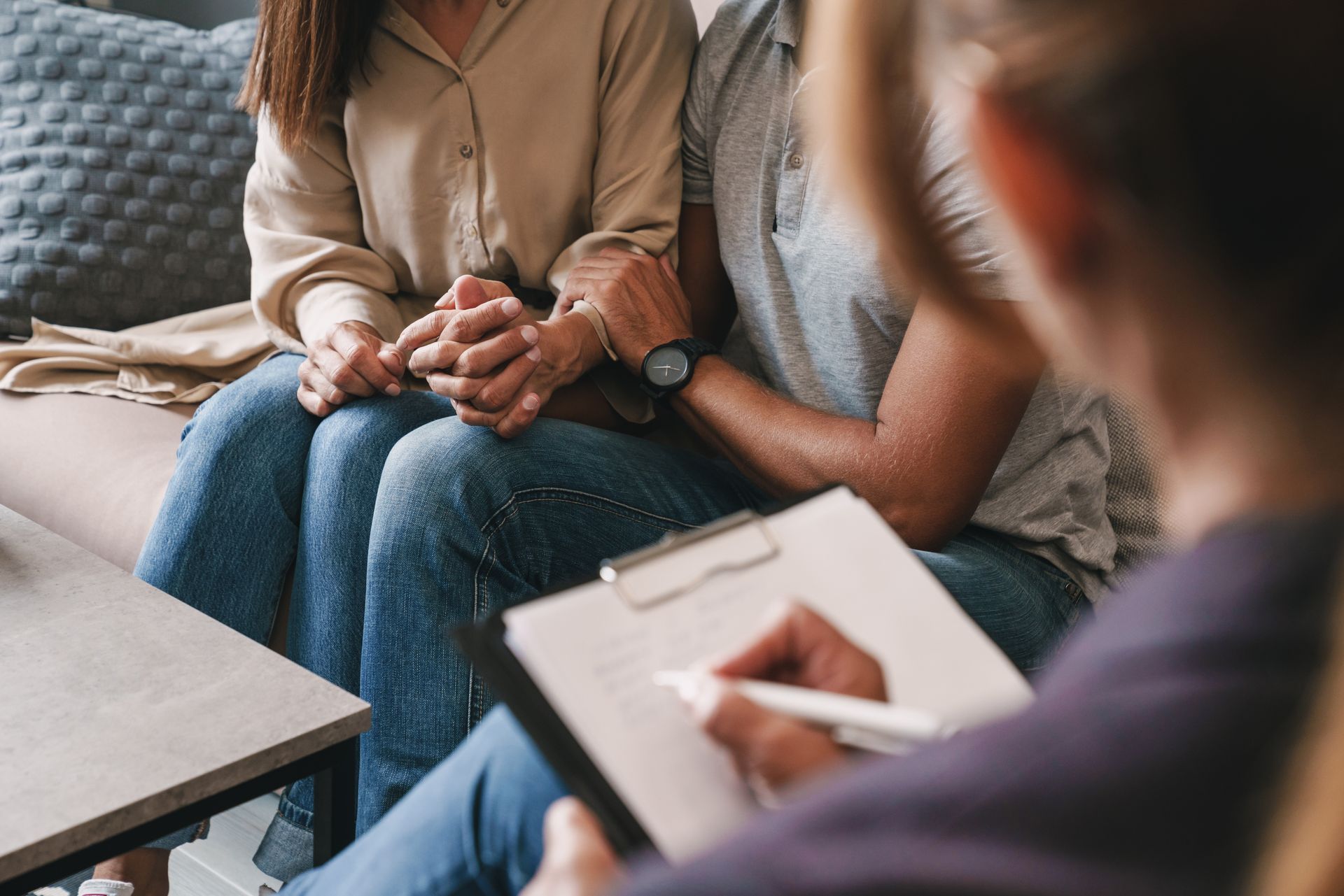 A man and a woman are sitting on a couch holding hands while a woman writes on a clipboard.