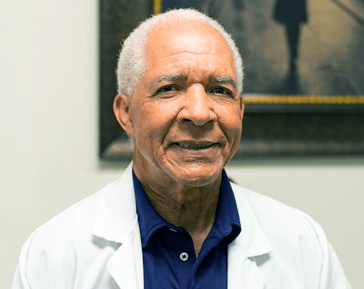 Dr. Symonett wearing a white lab coat and a blue shirt is smiling.