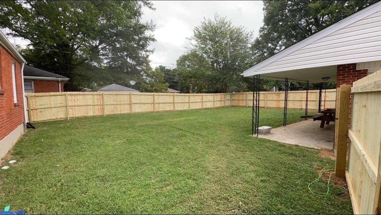 Backyard with grass lawn, tan wooden fence, red brick house, and covered patio on the right.