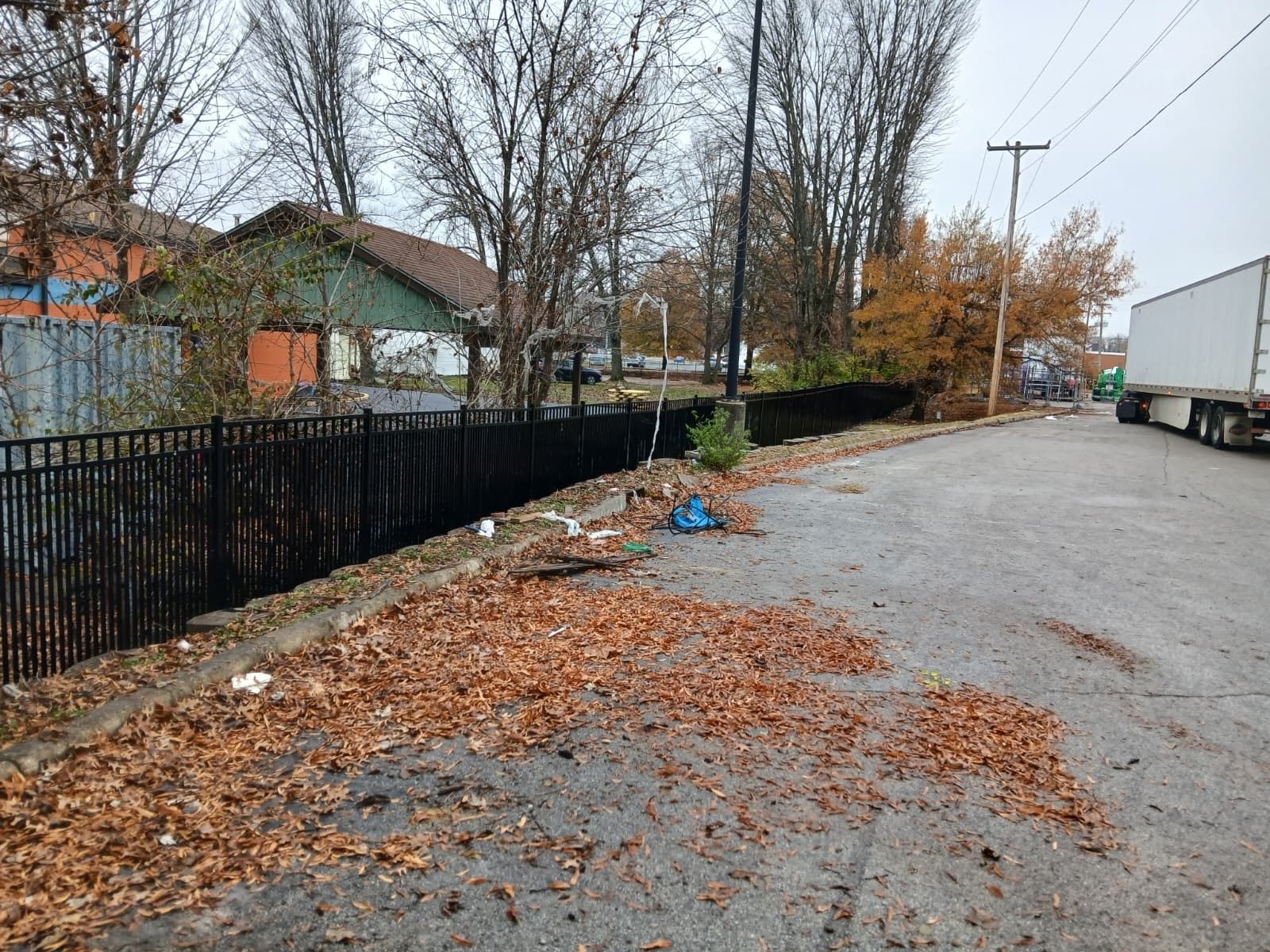 A black fence lines a littered road with fallen leaves. Buildings and a parked semi-truck are in the background.