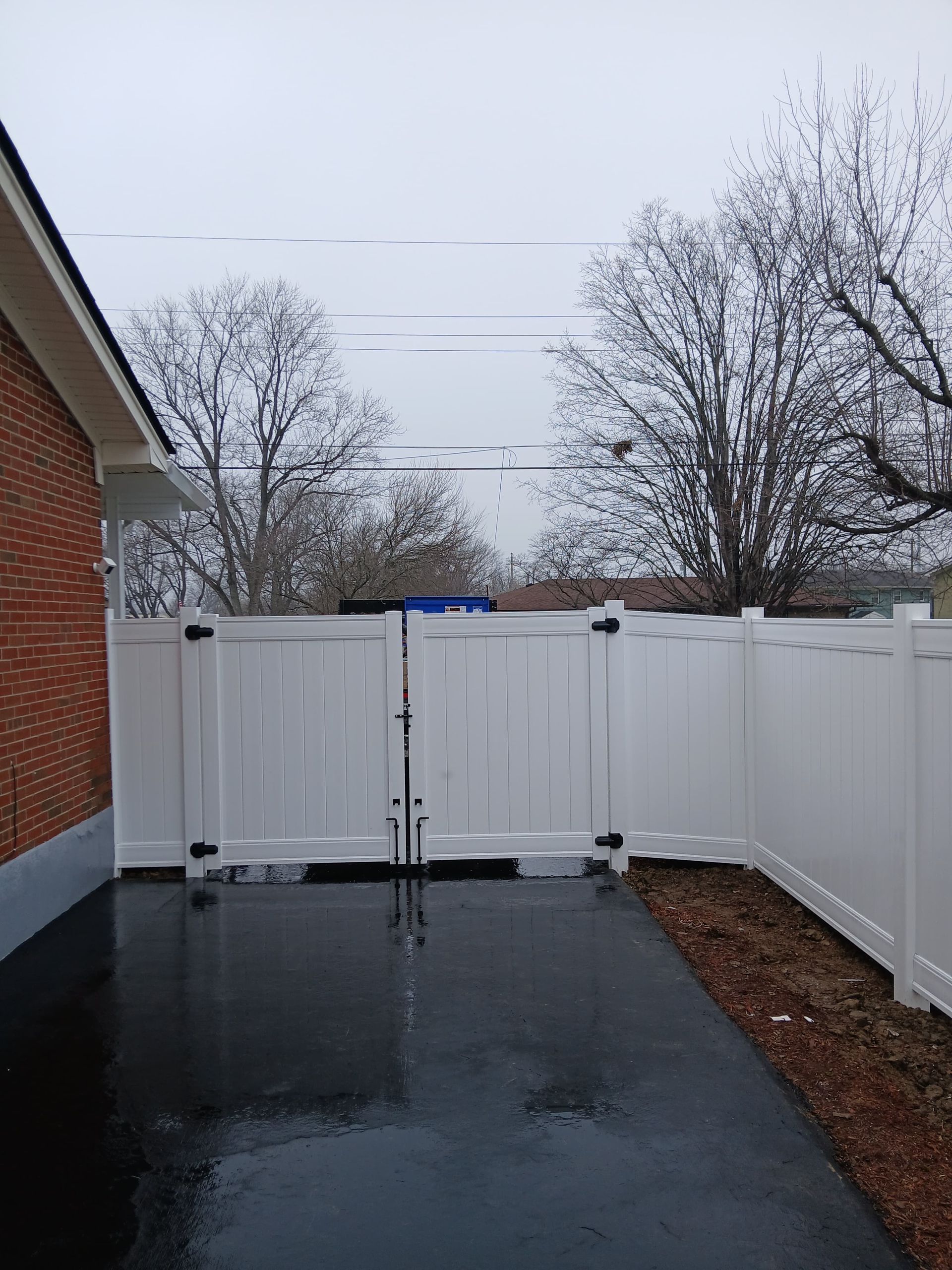 White vinyl fence with gate in front of a brick building. Black pavement and gray sky.