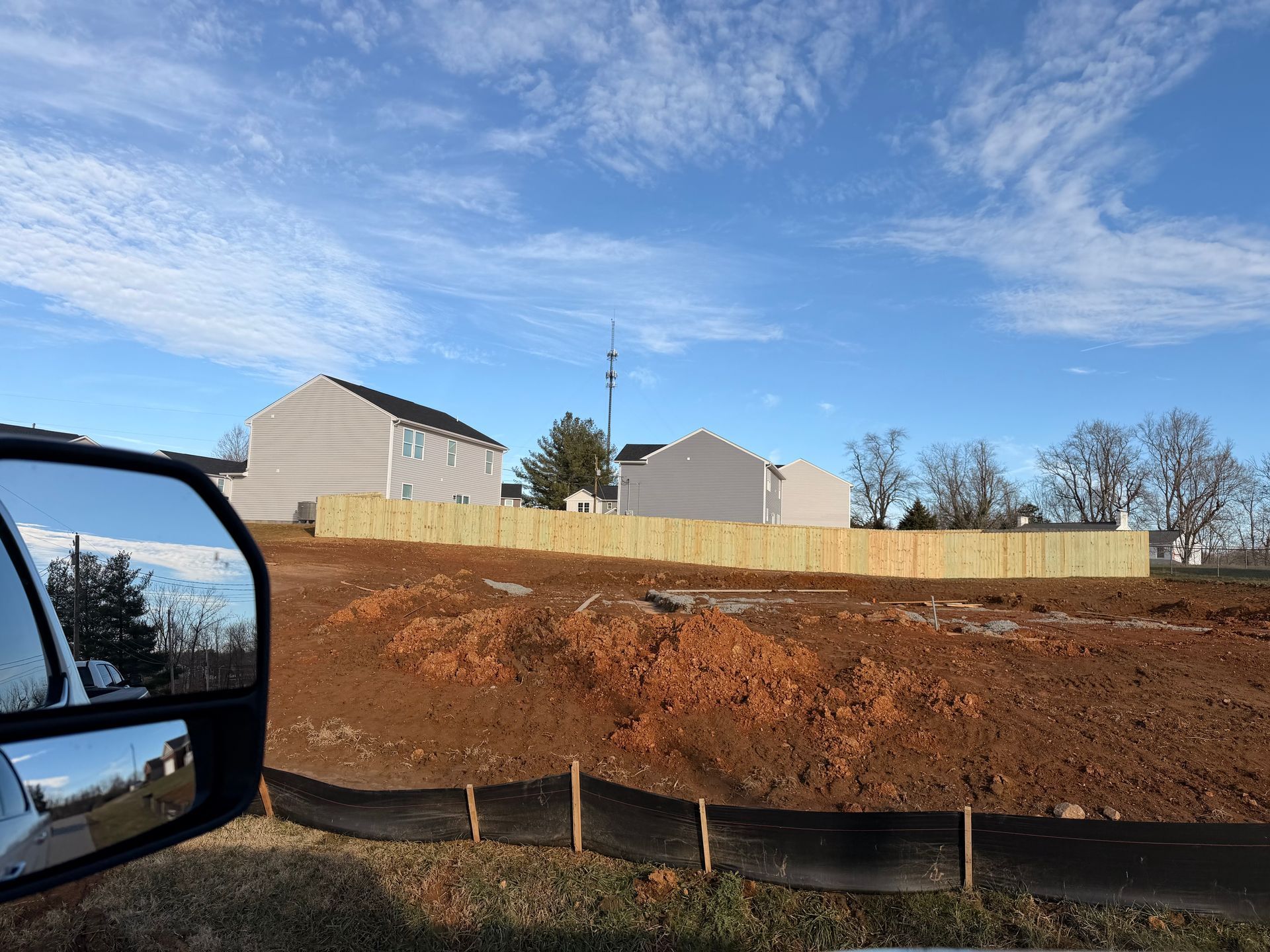 Cleared construction site with dirt, wooden fence, houses, and bright blue sky. Car mirror in foreground.