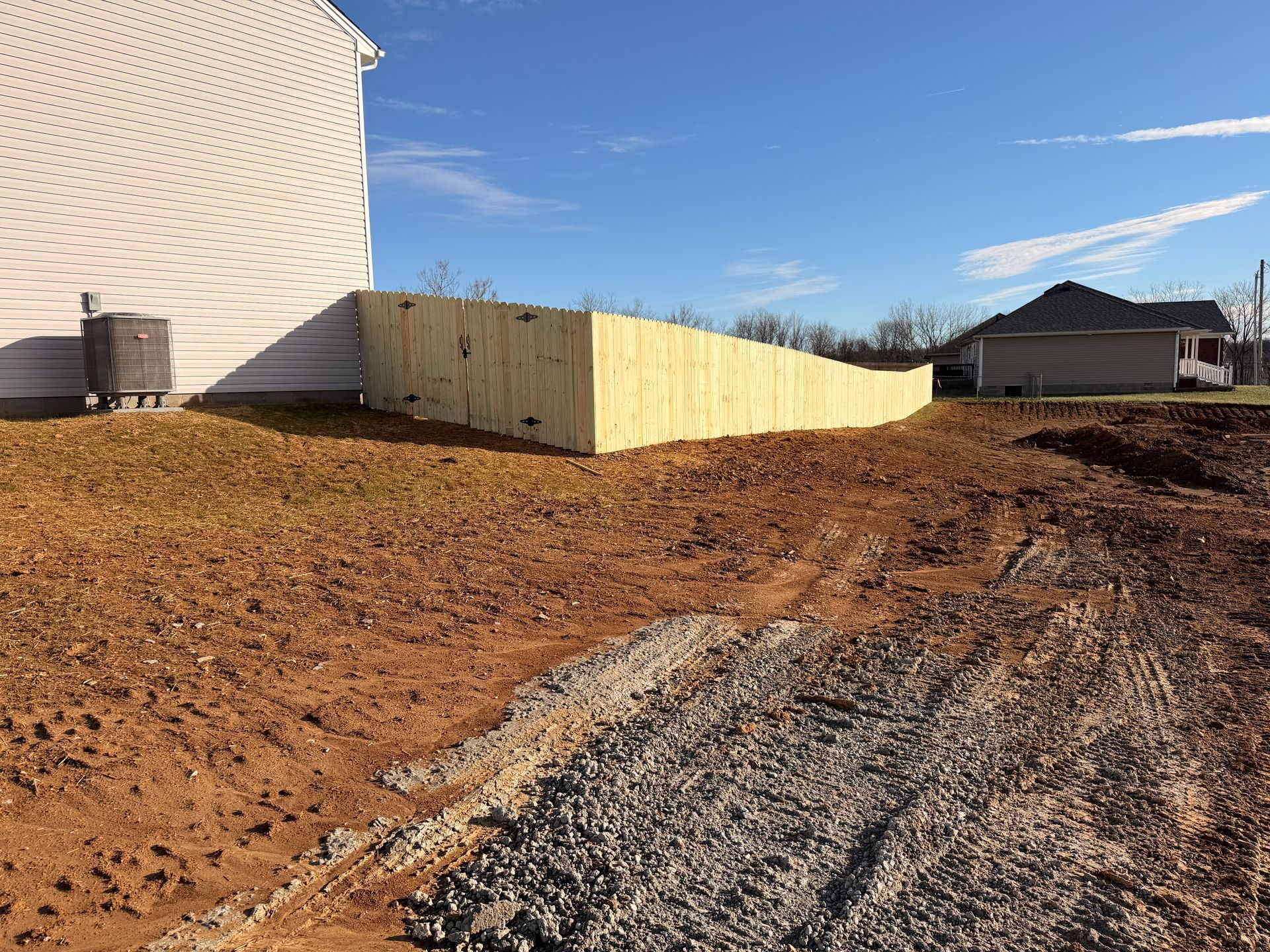 Wooden fence along a house, built on a slope of dirt and grass under a blue sky.