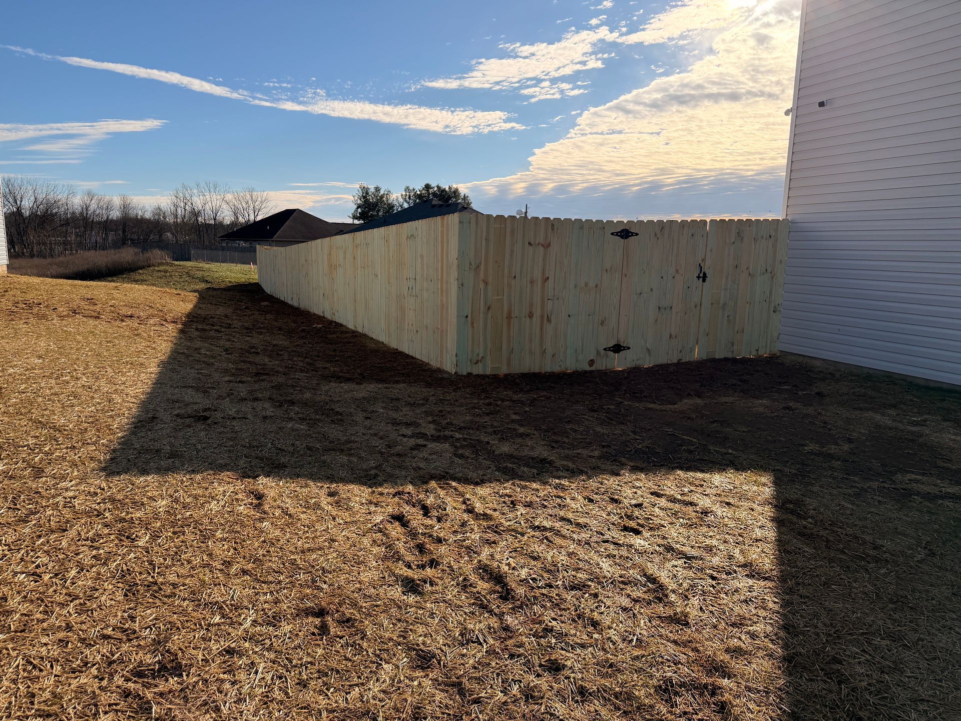 Wooden fence bordering a yard with mulch, sunny sky, and a white building.
