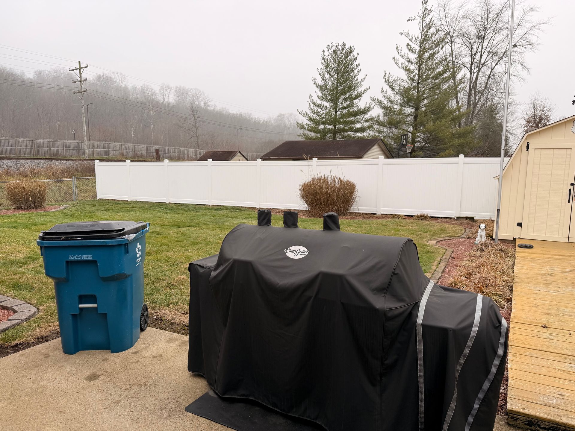 Backyard scene: covered smoker, blue trash can, white fence, and overcast sky.