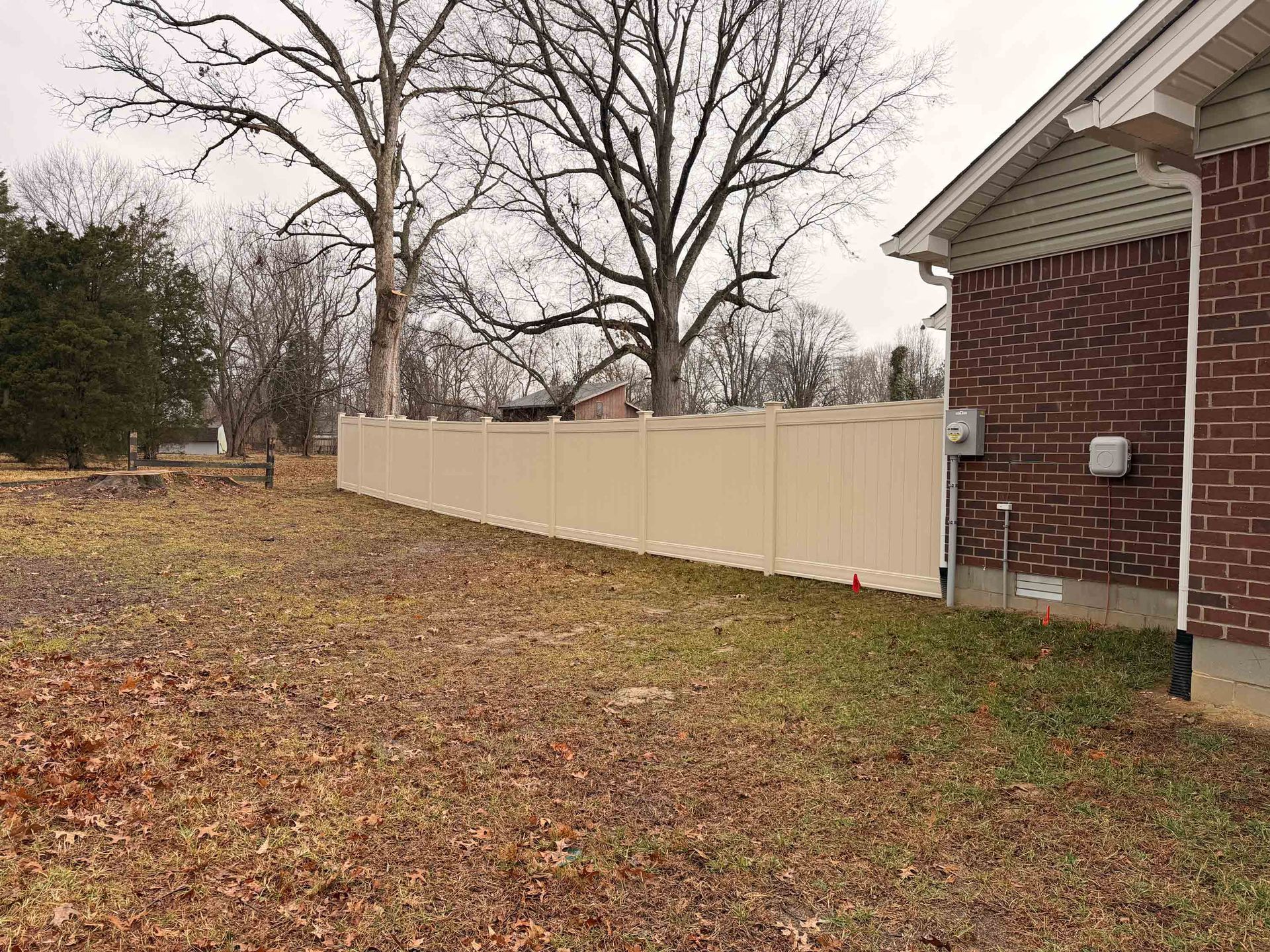 Beige fence along a yard with bare trees, brown grass, and a brick house on a cloudy day.