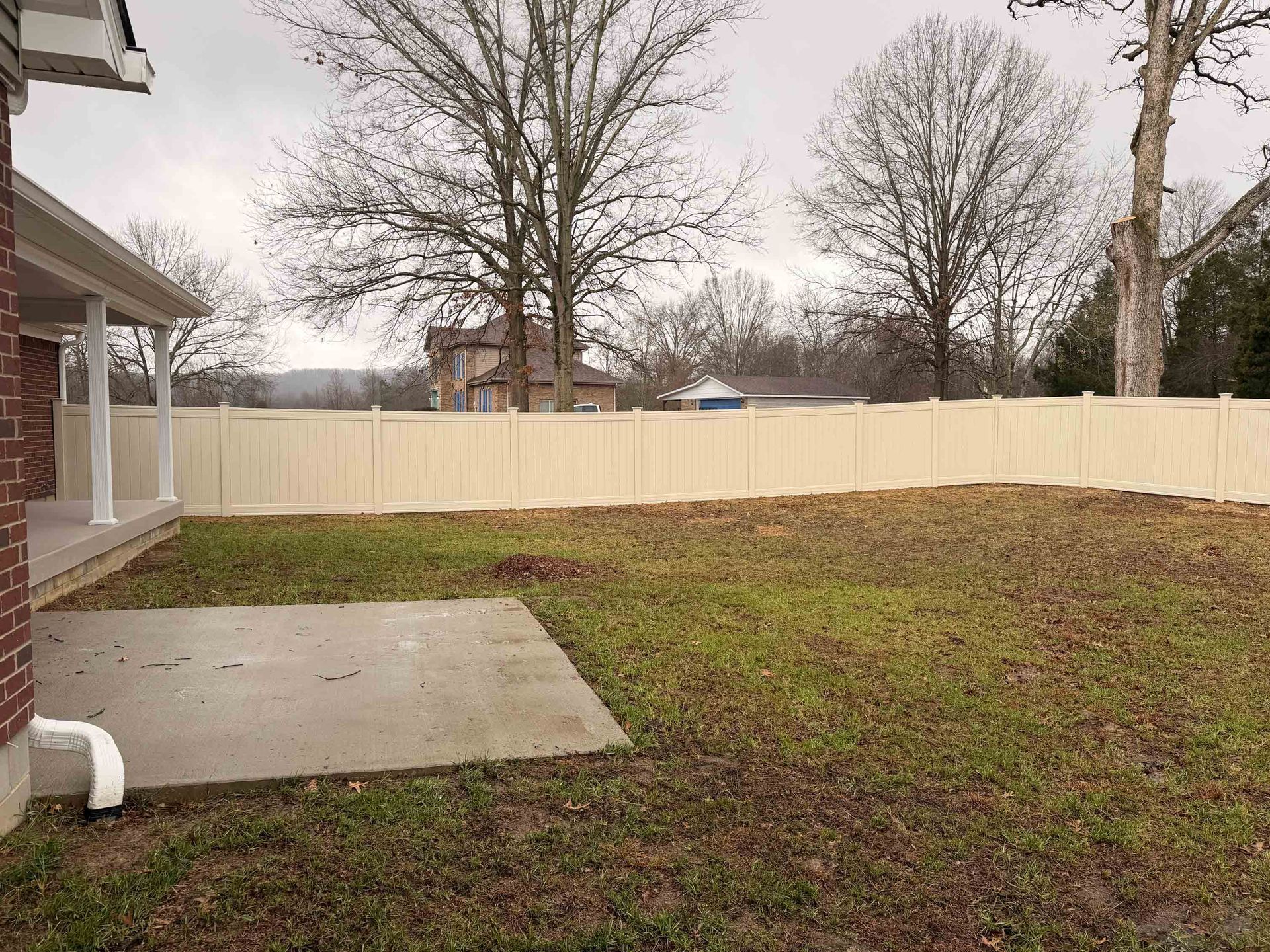 Backyard with beige fence, concrete patio, and bare trees under a cloudy sky.