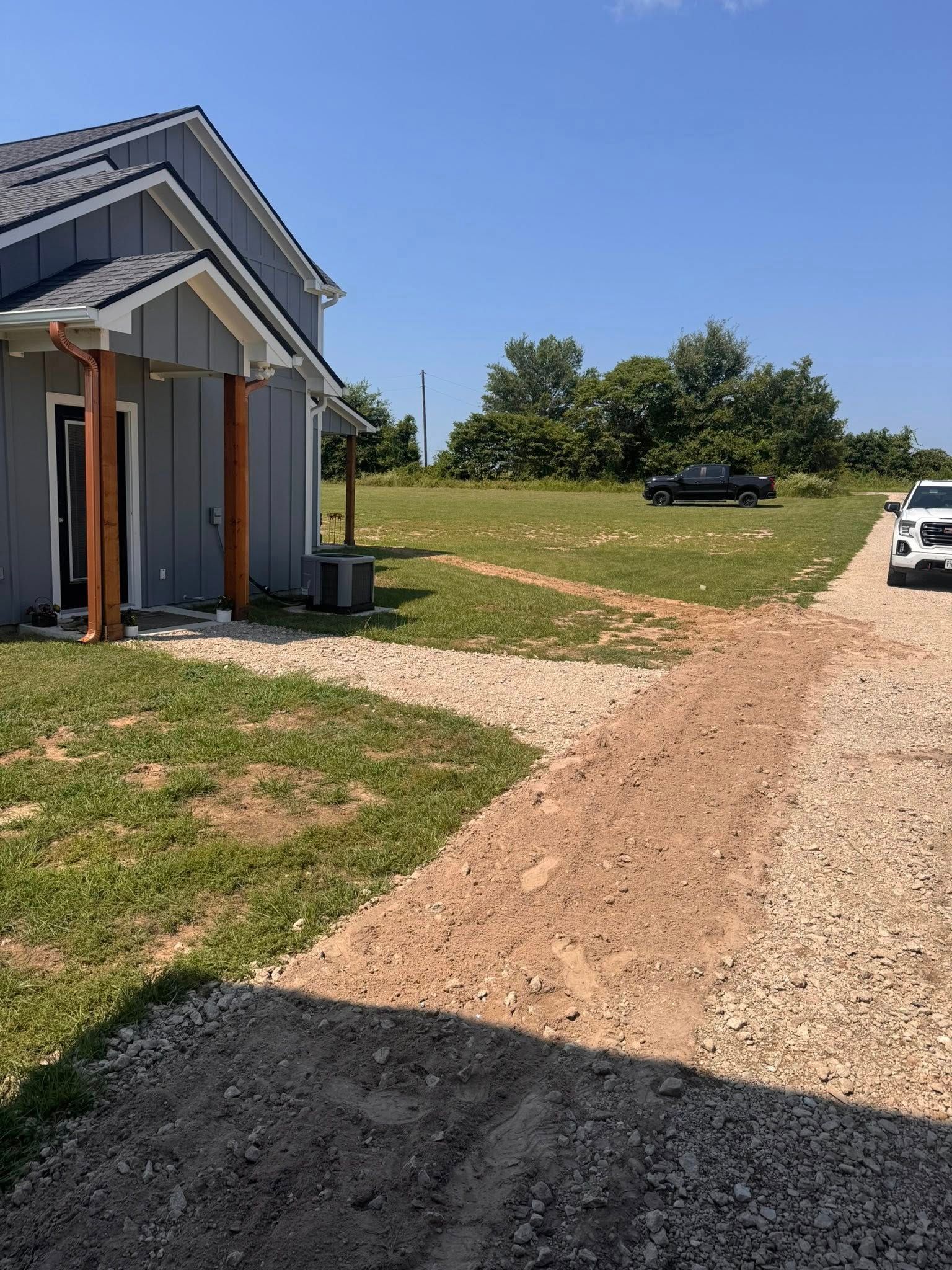 Gravel driveway next to a gray house with a brown front door on a sunny day. A car sits further away.