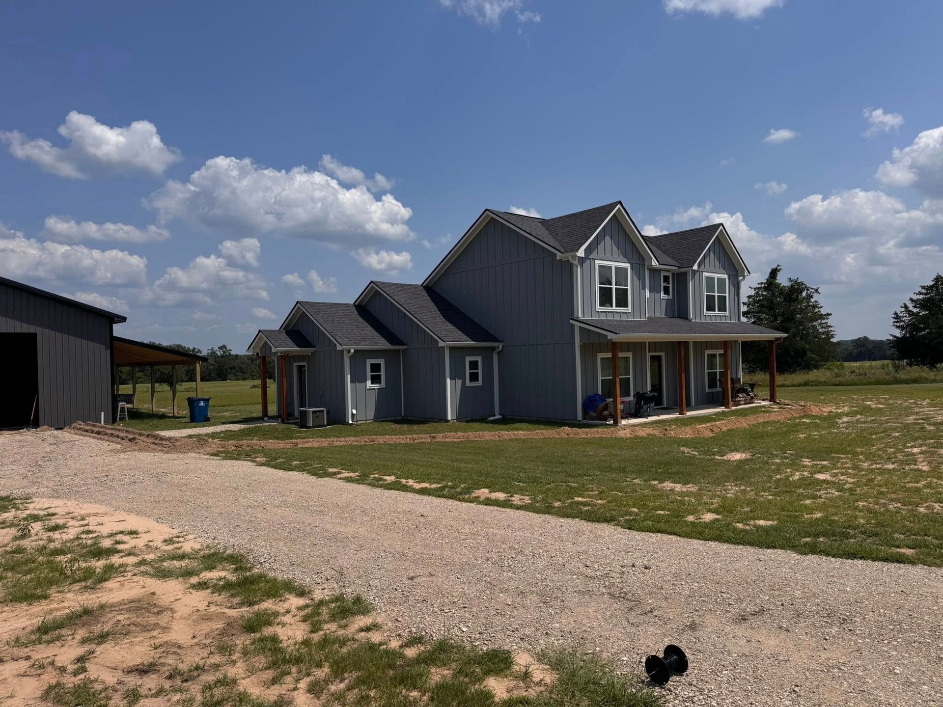 A two-story gray farmhouse with a covered porch, gravel driveway, and barn under a blue sky.