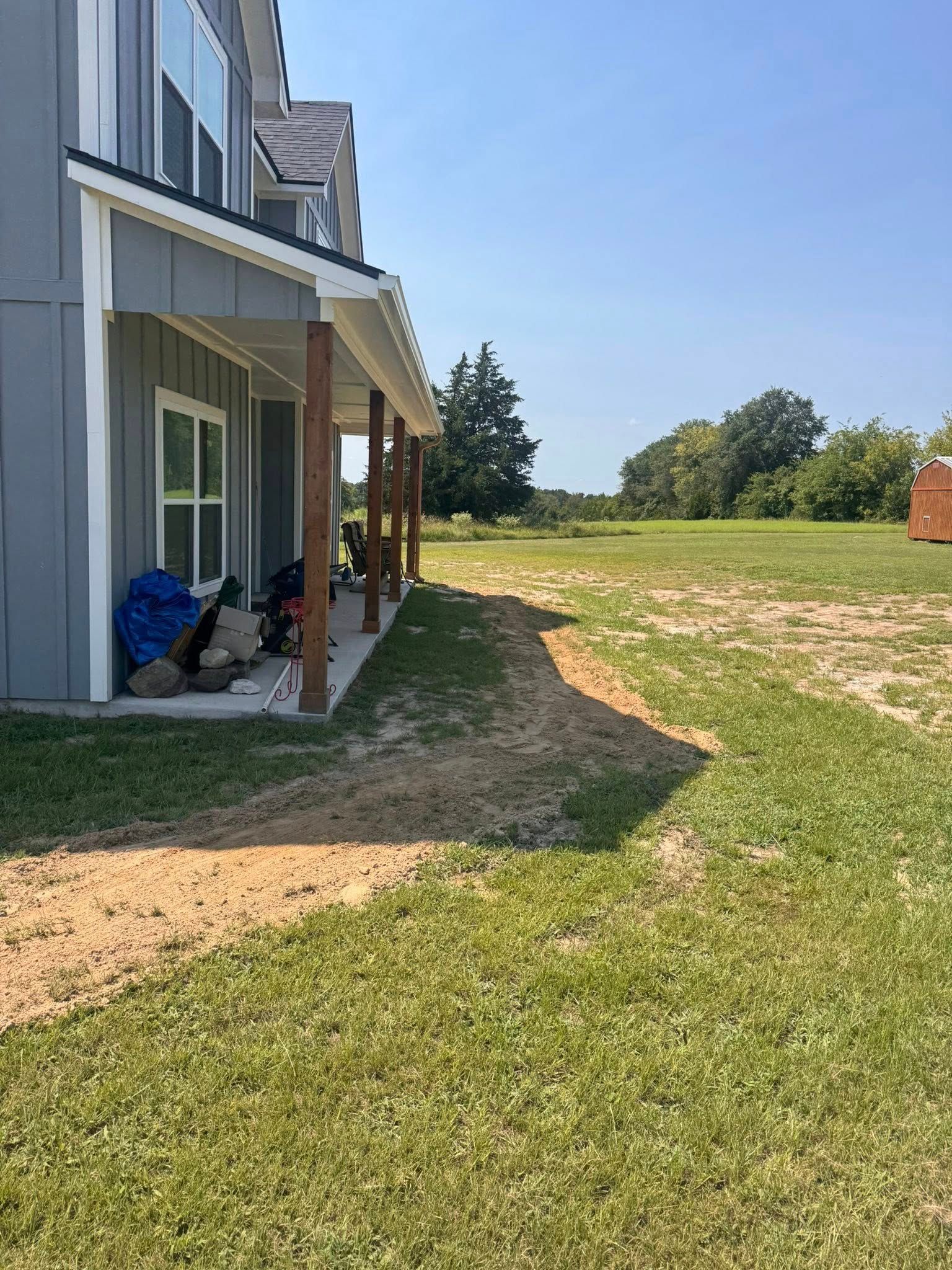 Side of a gray house with a porch, wooden beams, and a grassy yard on a sunny day.