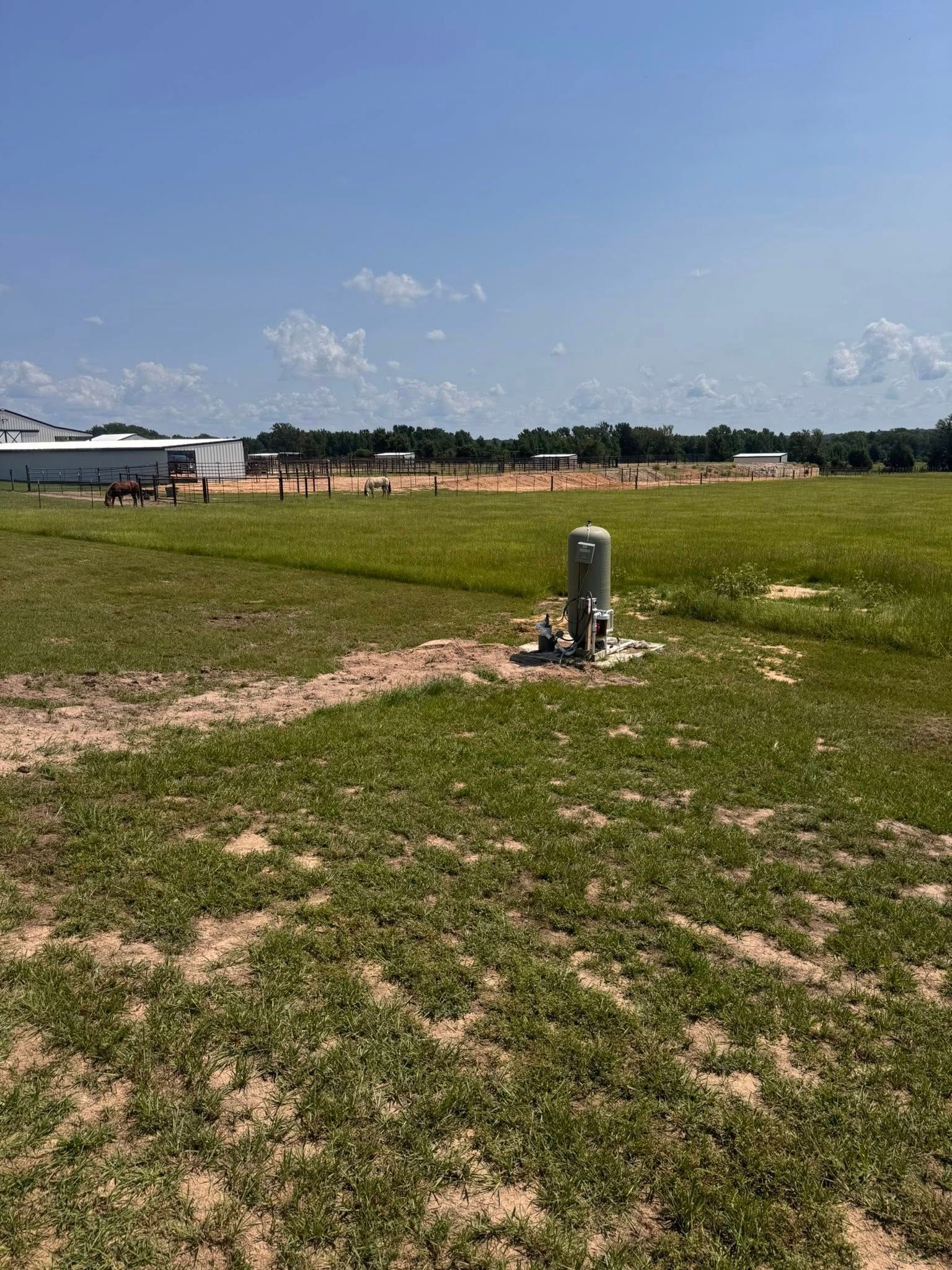 Grassy field with a well in the foreground, buildings and trees in the distance under a blue sky.