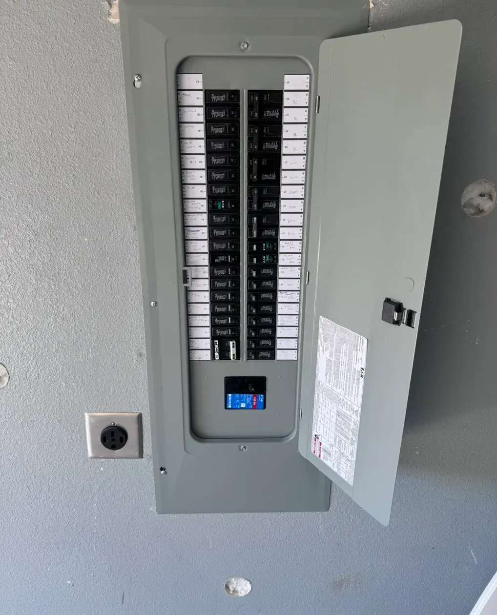 Gray electrical panel with open door, mounted on a textured wall, and a nearby outlet.
