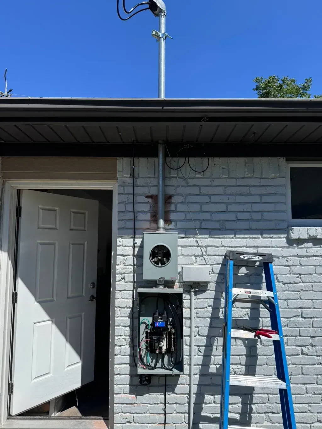 Electrical panel on brick building exterior; open door, blue ladder, conduit, wires, and sky.