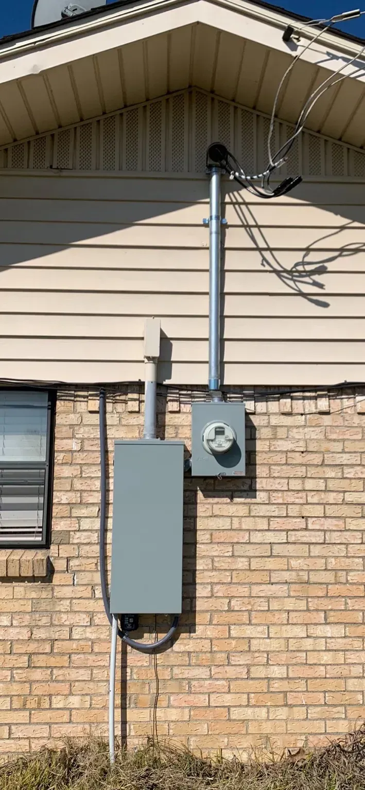 Electrical meter and service panel mounted on a brick wall with beige siding above. Wires run to the roof.