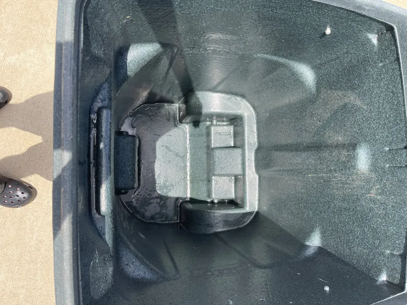 Top-down view of a dark trash can lid with a rectangular handle and textured surface.