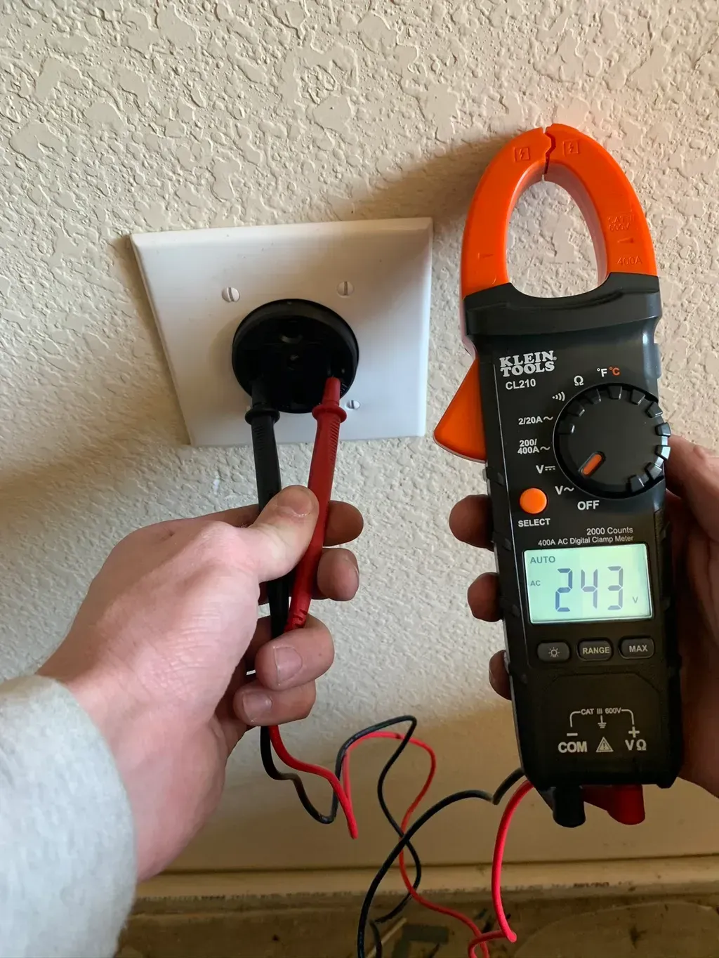 Hands testing an electrical outlet with a multimeter, which reads 243. The setting appears to be indoors.