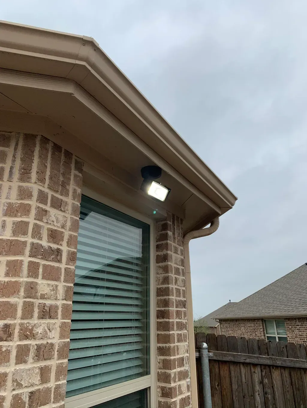 Exterior security light illuminates a brick wall, window, and gutter under a cloudy sky.
