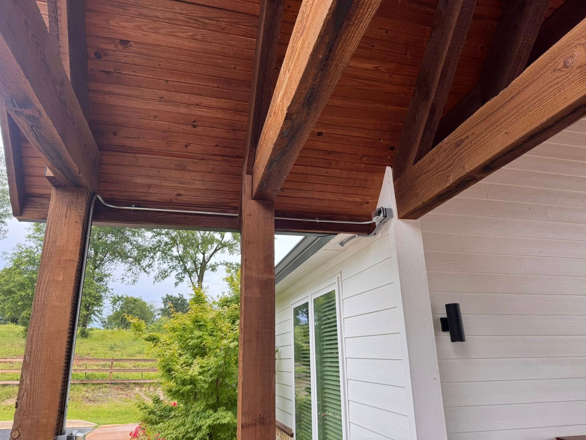 Wooden porch roof with supporting beams, alongside a white house exterior, overlooking a green landscape.