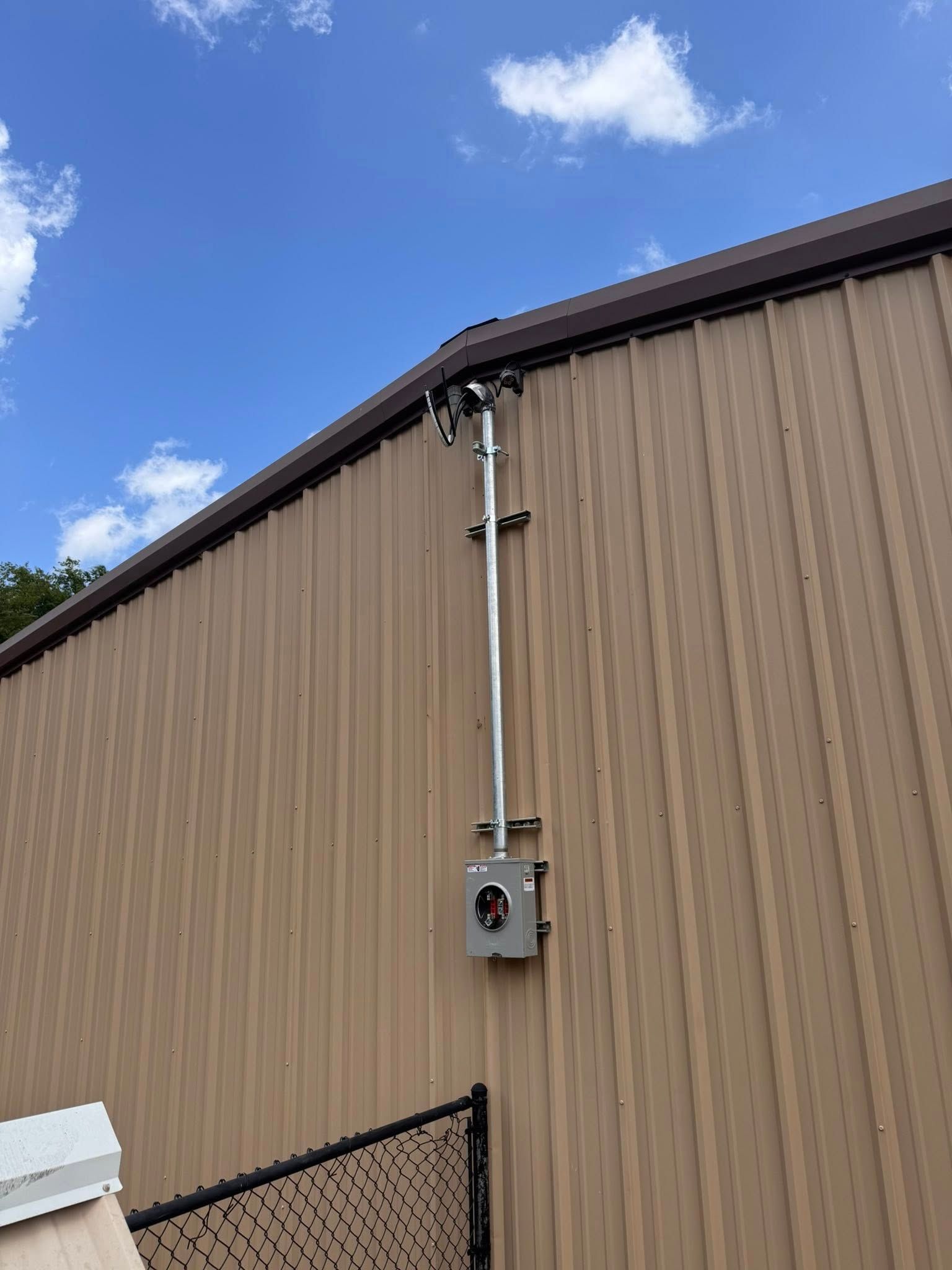 Electrical conduit and meter box mounted on the side of a brown metal building under a partly cloudy sky.