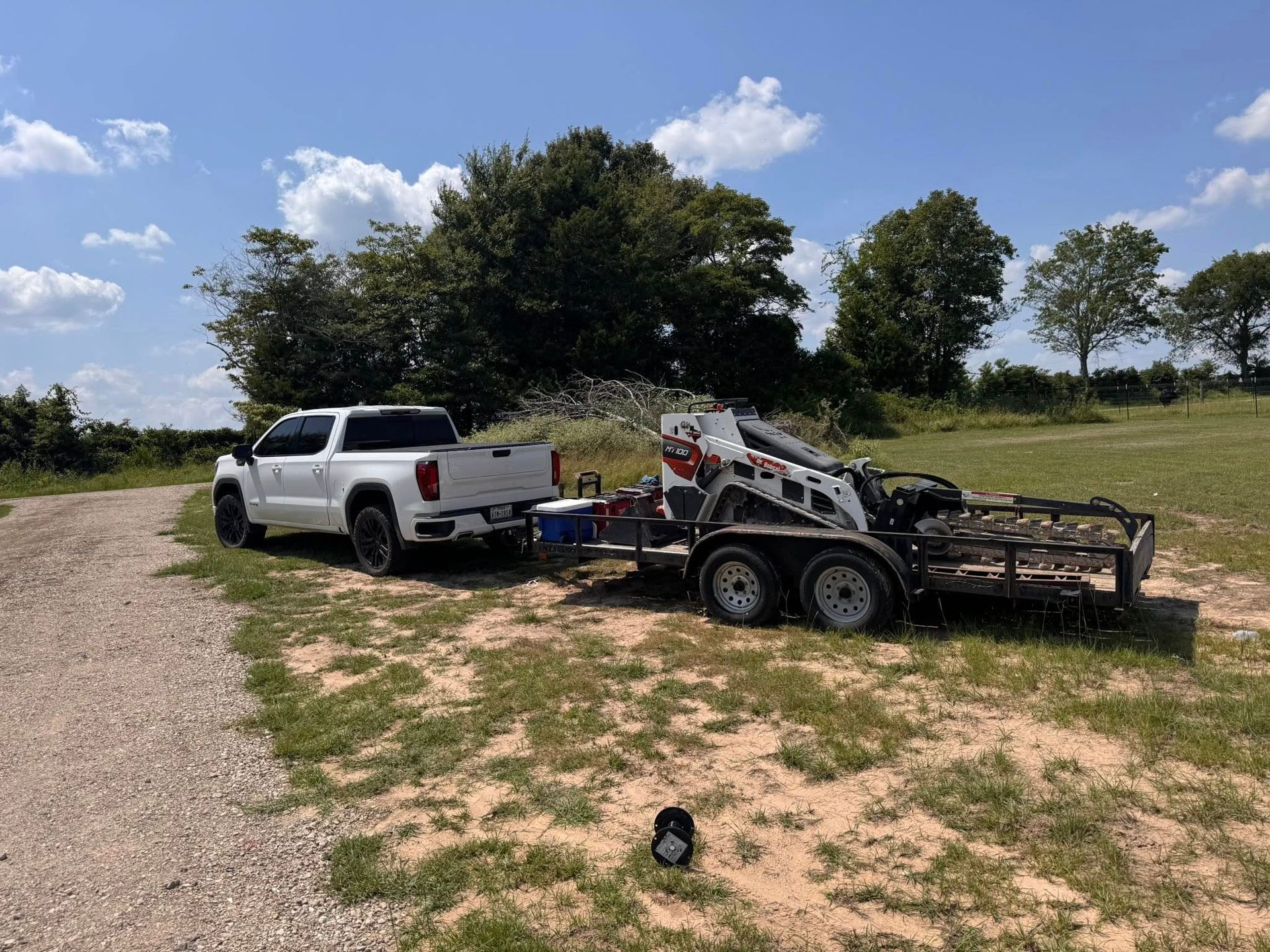 White pickup truck towing a trailer with a Bobcat and debris on a grassy field under a blue sky.