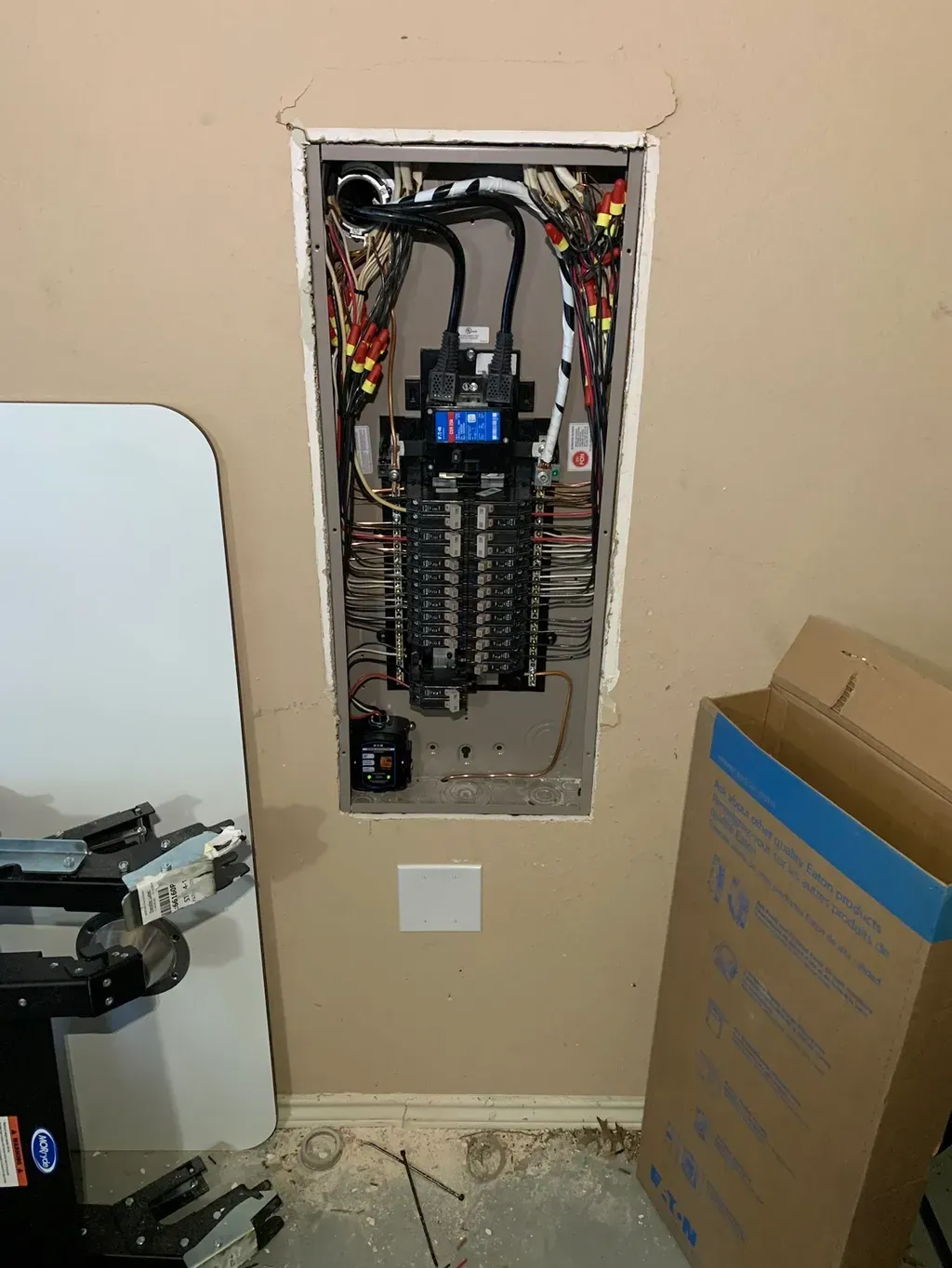 Electrical panel installed in a wall, with wires, breakers, and a white square outlet below it.