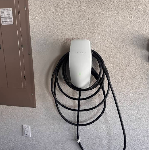 Tesla charging station mounted on a beige wall with the cable coiled below. Next to an electrical panel.
