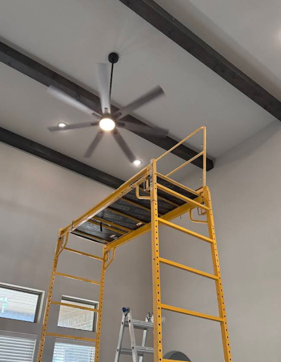 A yellow scaffold supports access to a ceiling fan, under dark beams, with windows below.