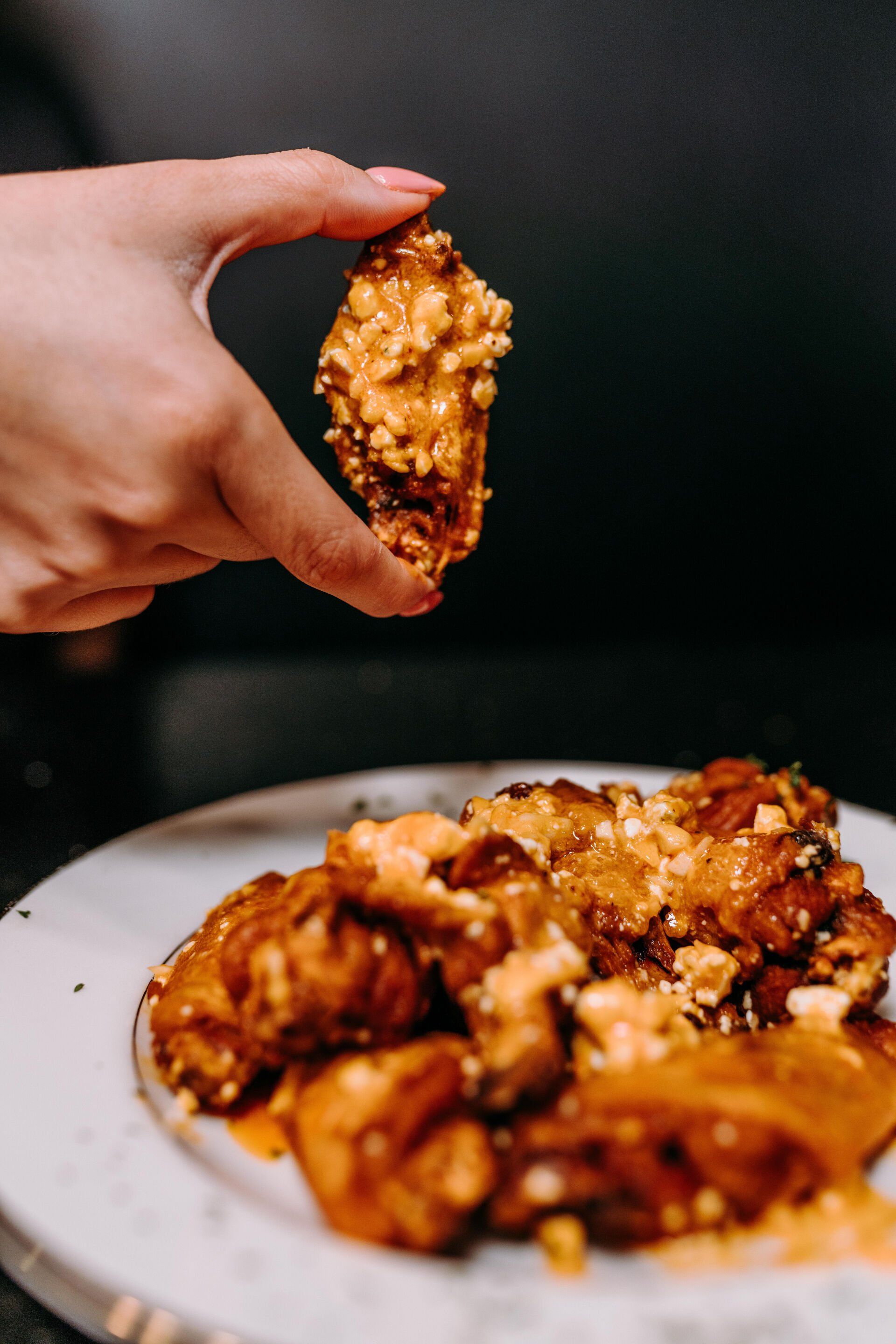 A person is holding a piece of chicken wing over a plate of chicken wings.