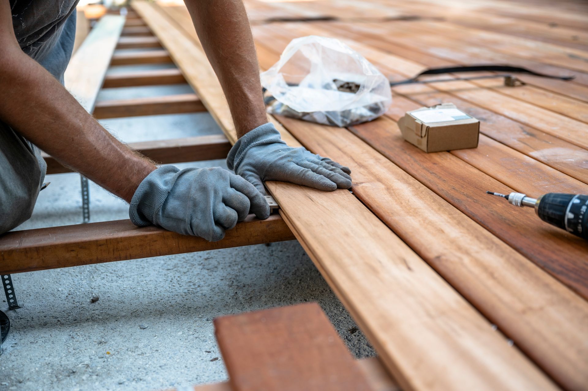 Person wearing gloves installing wooden deck planks outdoors. Tools and fasteners are nearby.
