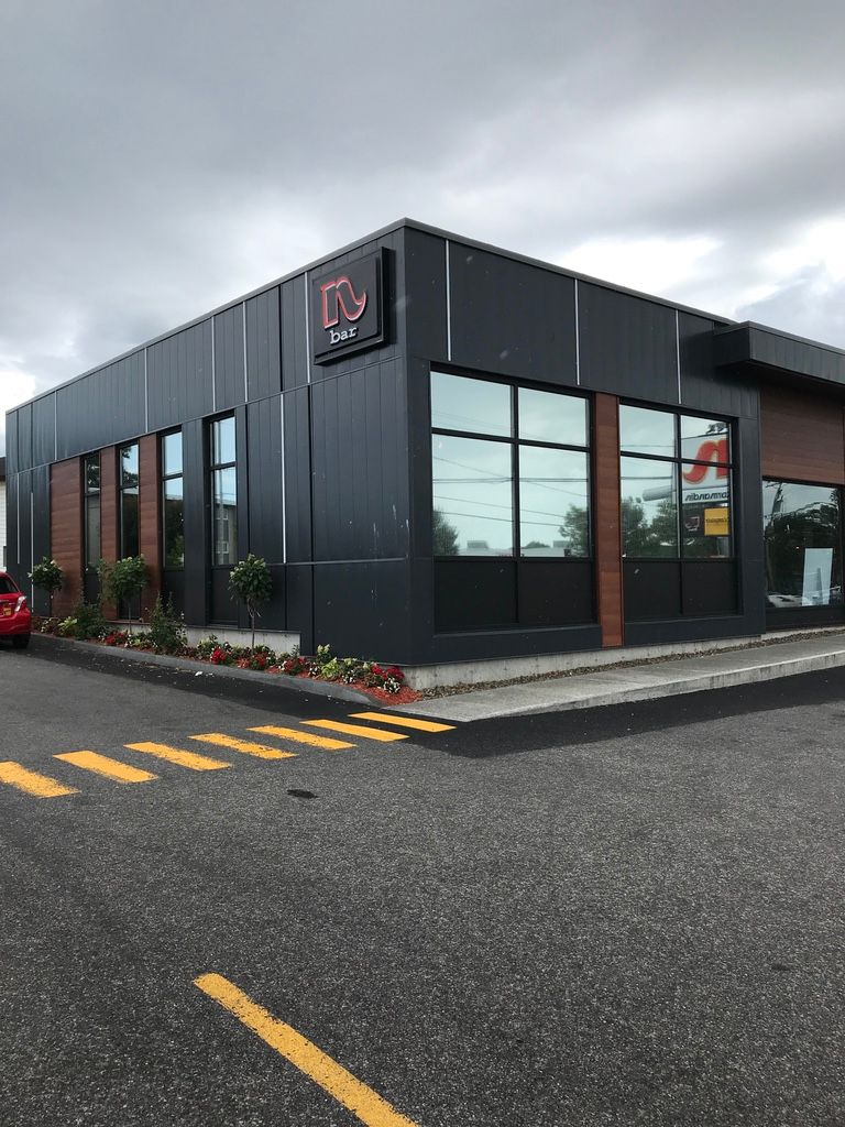 Bâtiment moderne, gris foncé avec de grandes fenêtres et une enseigne rouge, petits arbres devant, situé sur une route asphaltée.