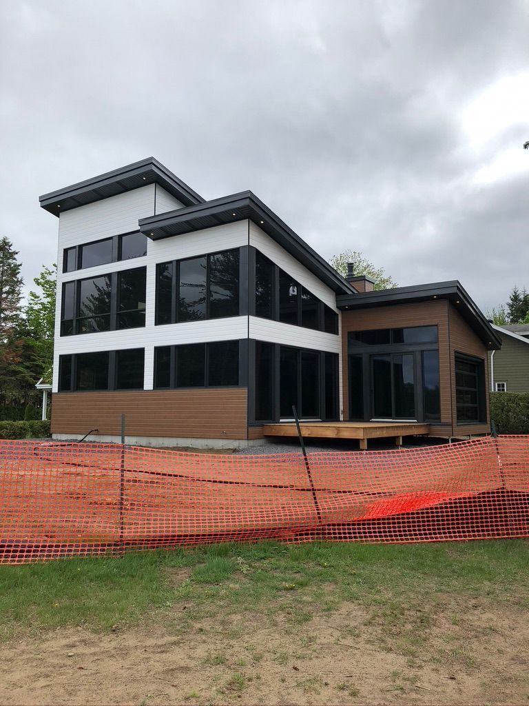 Maison moderne en construction; revêtement blanc et marron, fenêtres foncées, clôture de construction rouge.