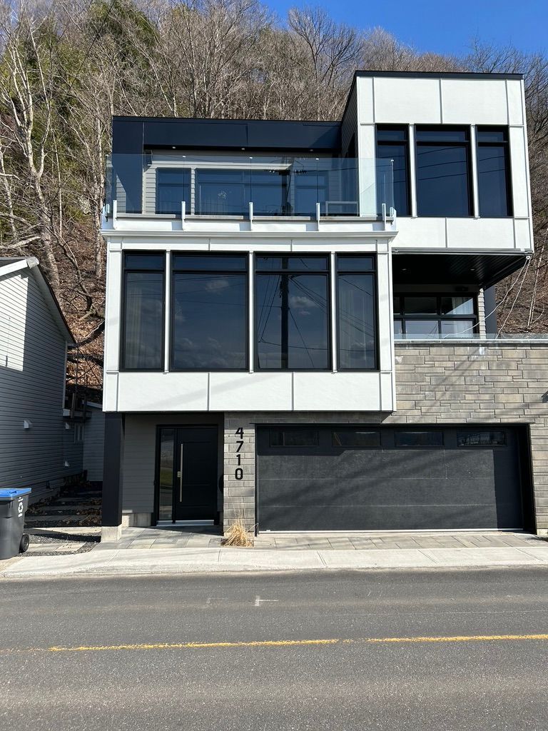 Maison moderne à étages avec boiseries et fenêtres noires et garage. Façade en pierre et blanche. Balcon avec garde-corps en verre.