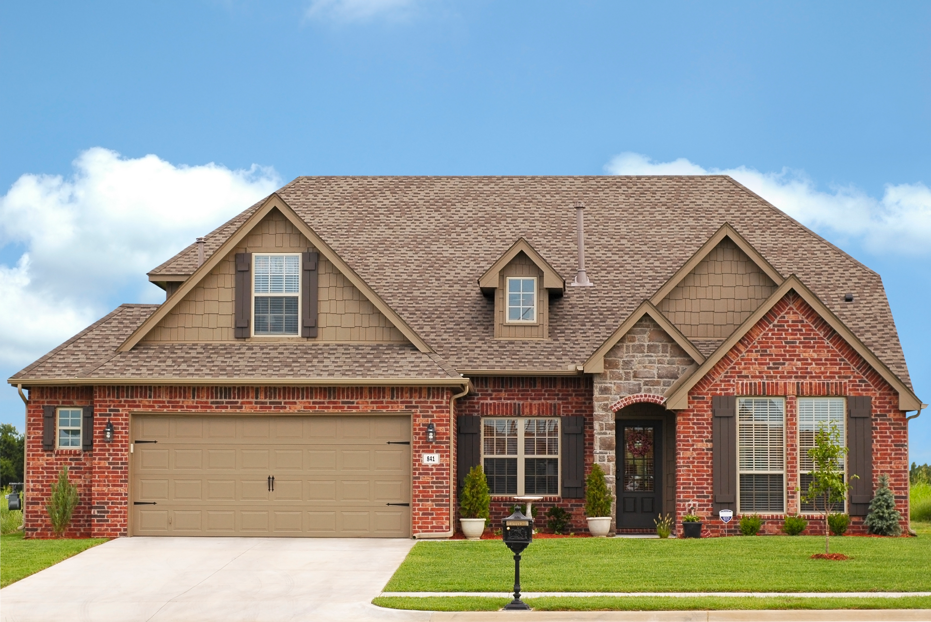 Red brick house with a tan garage door and brown roof against a blue sky.