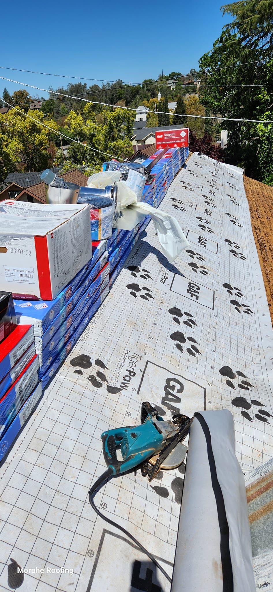 A roof being worked on with supplies and a tool visible against a background of trees and blue sky.