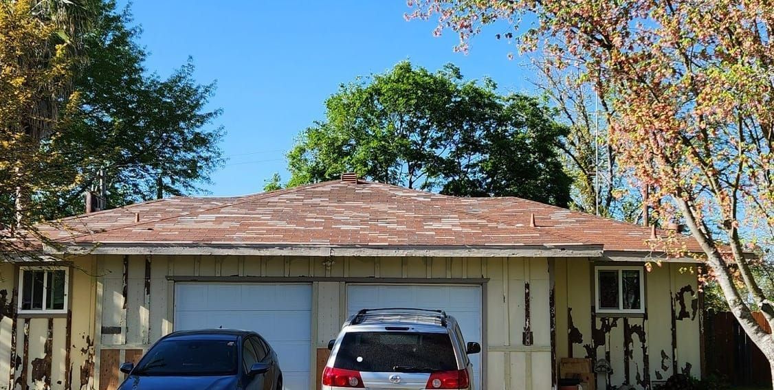 Garage with peeling paint, two vehicles parked inside, trees and blue sky above.
