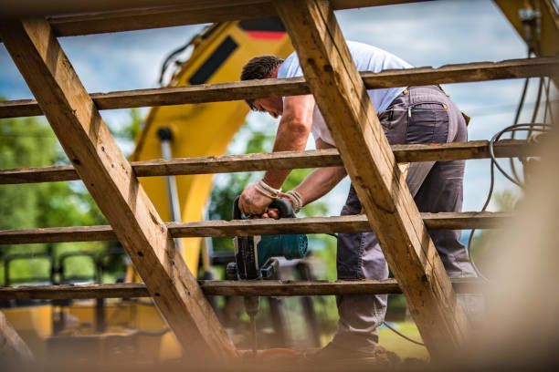 Construction worker using a drill on a wooden structure, with a yellow excavator in the background.